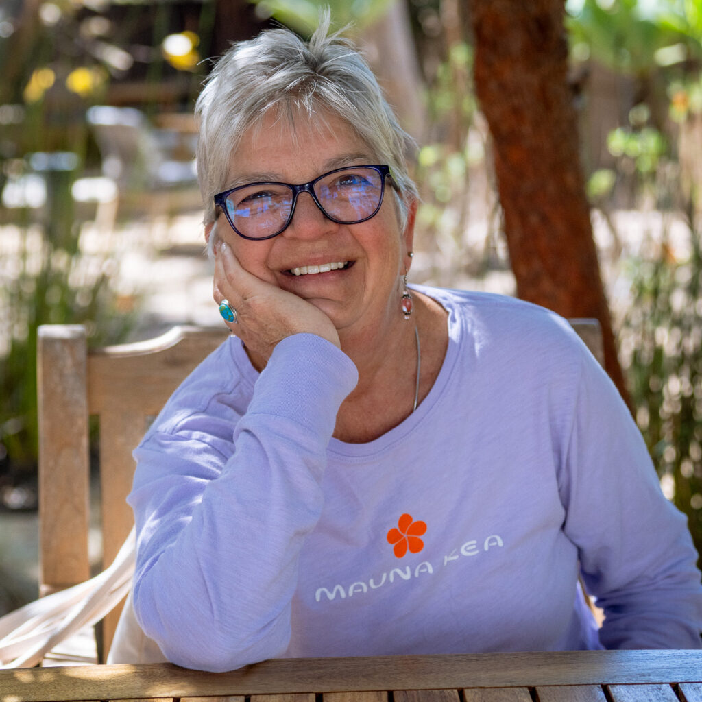 Smiling older woman with short gray hair and glasses sits at a wooden table outdoors, resting her head on her hand, wearing a light purple “Mauna Kea” shirt. Sunlight, greenery, and andre J-inspired style shine through.