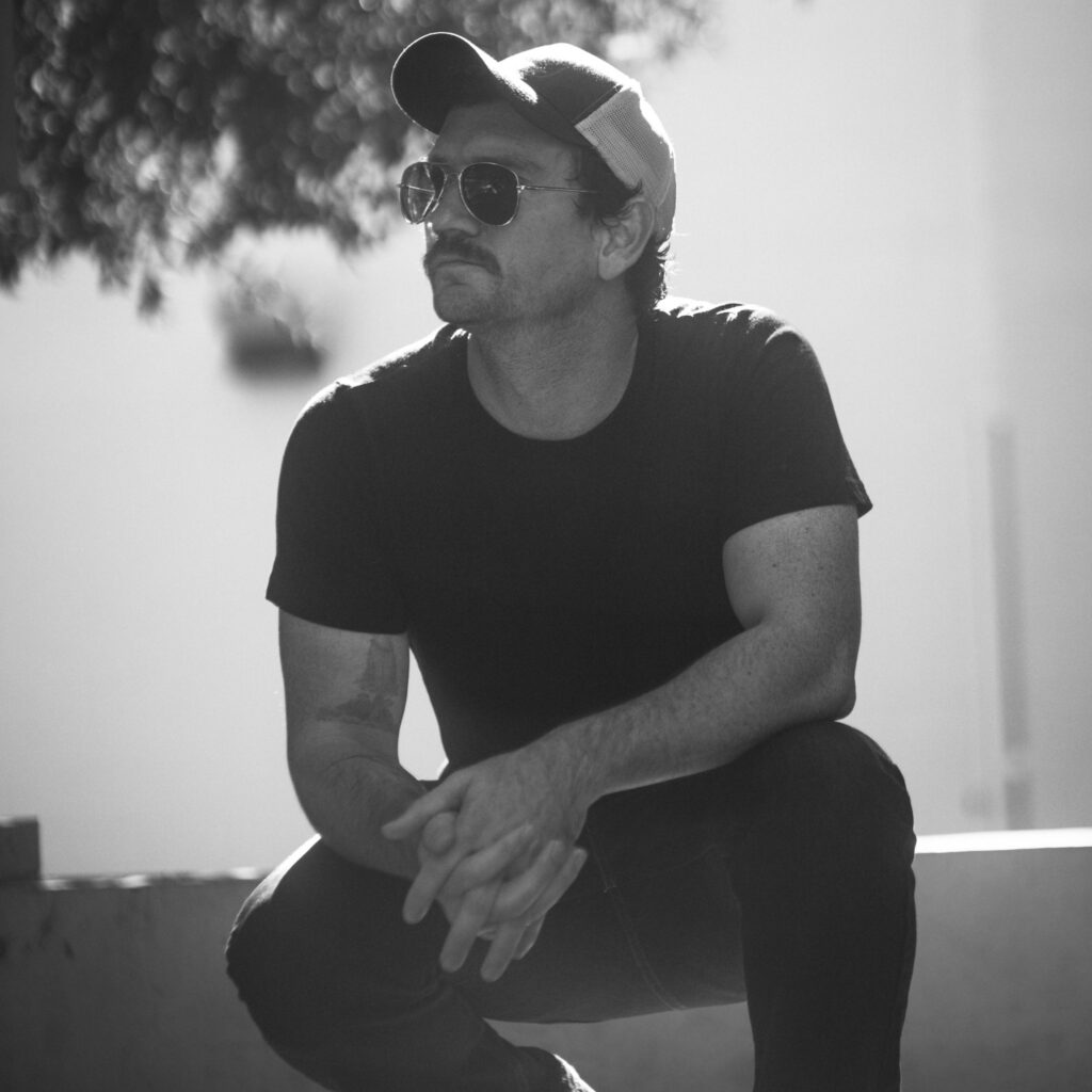 A man with a mustache, wearing a baseball cap and sunglasses, crouching with his hands clasped.